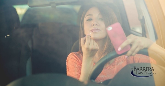 Girl in her car holding onto the steering wheel and her phone with one hand and applying lipstick with the other hand.